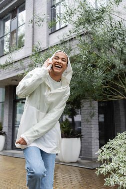 Kapüşonlu su geçirmez yağmurluk giymiş, yağmurun altında dururken akıllı telefondan konuşan neşeli Afro-Amerikan kadını. 