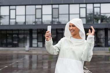 Su geçirmez yağmurluk giyen mutlu Afro-Amerikalı kadın yağmurluk sırasında akıllı telefondan selfie çekiyor. 