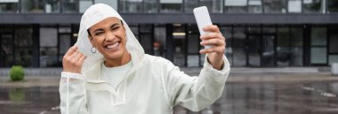 cheerful african american woman in waterproof raincoat taking selfie on smartphone during rain, banner  clipart