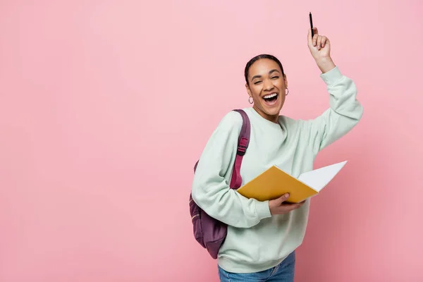 excited african american student holding notebook and pen while standing with backpack isolated on pink