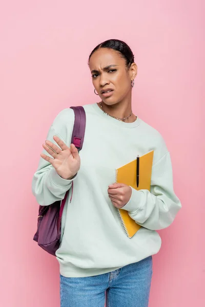 stock image displeased african american student holding notebook and pen while showing refuse gesture isolated on pink 