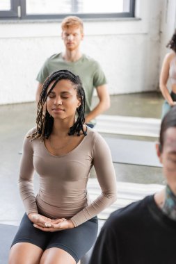 Afrika kökenli Amerikalı kadın yoga dersinde bulanık insanların yanında meditasyon yapıyor. 