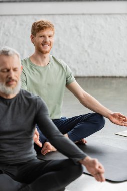 Neşeli kızıl saçlı adam gyan mudra yapıyor ve yoga stüdyosunda kameraya bakıyor. 