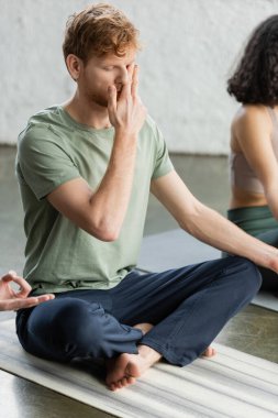 Genç kızıl saçlı adam yoga stüdyosunda burun deliği nefes alma alıştırması yapıyor. 