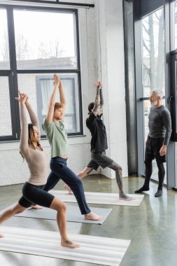 Crescent Lunge Asana 'da yoga dersinde olgun koçun yanında duran ırklar arası insanlar. 