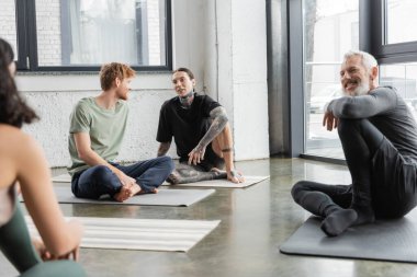 Yoga dersinde minderlerin üzerinde oturan genç erkekler konuşuyor. 