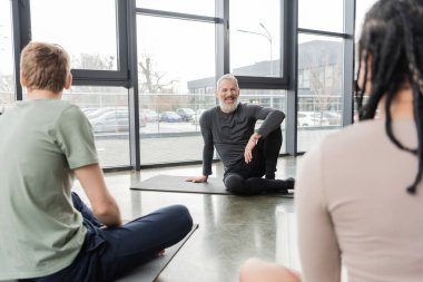 Positive coach looking at blurred multiethnic people in yoga studio  clipart