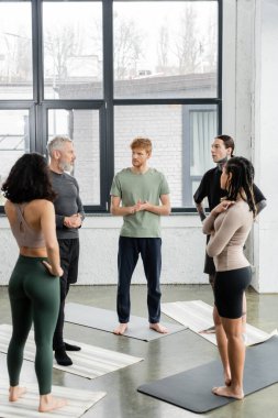 Interracial people looking at mature coach while standing on mats in yoga class  clipart