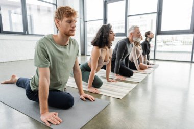 Kızıl saçlı adam yoga sınıfında çok ırklı insanların yanında Half Pigeon Asana egzersizi yapıyor. 