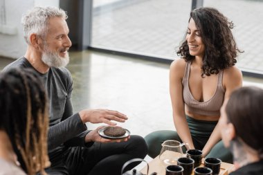 Gri sakallı dövmeli adam yoga stüdyosunda neşeli orta doğulu kadının yanında sıkıştırılmış puer çayı tutuyor. 