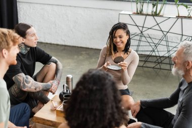 happy african american woman with dreadlocks holding compressed puer tea near people in yoga studio  clipart