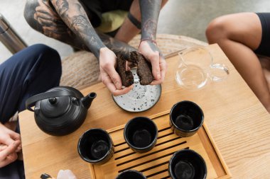 Geleneksel Çin çay demliği ve fincanlarının yanında sıkıştırılmış puer çayı kıran dövmeli adamın üst görüntüsü. 