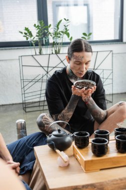 young man with tattoos smelling fermented puer tea near burning Palo Santo stick and Chinese teapot with cups  clipart