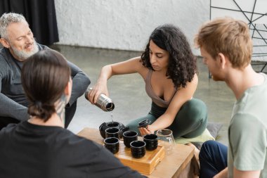 Kıvırcık orta doğulu kadın termostan sıcak su dolduruyor ve yoga stüdyosunda erkeklerin yanında puer çayı demliyor. 