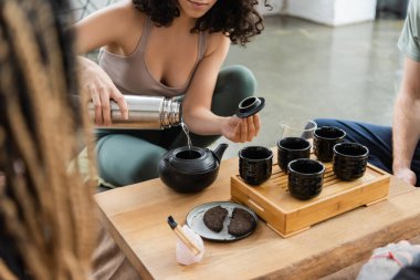 cropped view of woman pouring hot water from thermos while brewing pu-erh tea near people in yoga studio  clipart