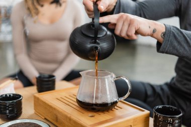 cropped view of tattooed man pouring freshly brewed puer tea into glass jug near woman on blurred background  clipart