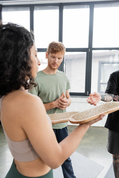 Redhead man standing near middle eastern woman with sadhu board in yoga class 