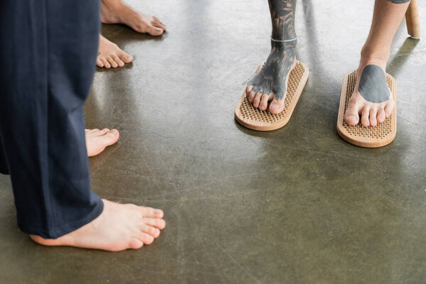 cropped view of tattooed man standing with bare feet on sadhu nail board near faceless people in yoga studio