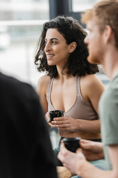 cheerful middle eastern woman with curly hair smiling during tea ceremony in yoga studio 