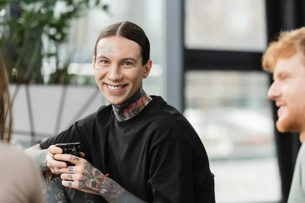 stock image tattooed man smiling while holding japanese cup with tea near people on blurred foreground 