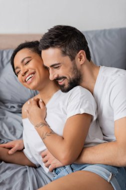 Memnuniyetle sakallı adam, güzel Afro-Amerikan kadınına sarılıyor. Evde gözleri kapalı gülümsüyor.
