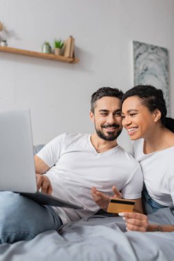 Evdeki yatak odasında online alışveriş yaparken kredi kartıyla Afrikalı Amerikalı bir kadının bilgisayarına bakan kaygısız bir adam.