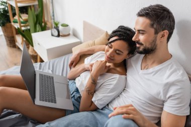 Beyaz tişörtlü kaygısız çift yatak odasındaki laptopta film izliyor.