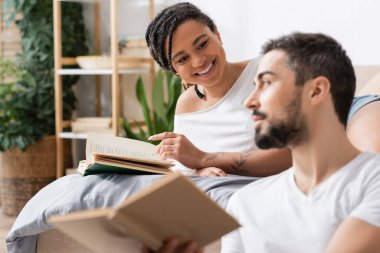 Neşeli Afro-Amerikalı kadın evde, yatak odasında bulanık erkek arkadaşının yanındaki kitaba işaret ediyor.