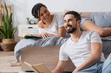 carefree bearded man sitting with book near young african american girlfriend lying on bed at home clipart