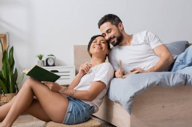 carefree african american woman with book smiling with closed eyes while sitting near smiling boyfriend lying on bed at home clipart