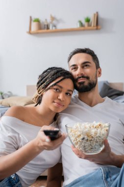 Elinde uzaktan kumandayla gülümseyen Afrikalı Amerikalı kadın evde yatak odasında bir kase patlamış mısırla sakallı erkek arkadaşının yanındaki televizyon kanallarını tıklatıyor.