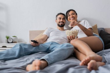 Memnuniyetle Afro-Amerikalı kadın evde yatağında televizyon kumandası olan sakallı, gülümseyen bir adamın yanında patlamış mısır yiyor.