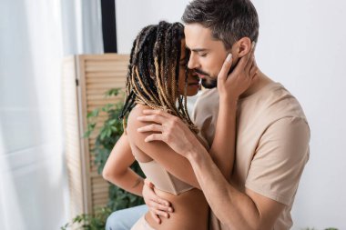 Tişörtlü sakallı adamın yatak odasında rastalı saçlarıyla genç ve seksi Afro-Amerikan kadınına sarılışının yan görüntüsü.