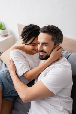 Genç Afrikalı Amerikalı kadın ve beyaz tişörtlü mutlu sakallı adam yatak odasında kucaklaşıyorlar.