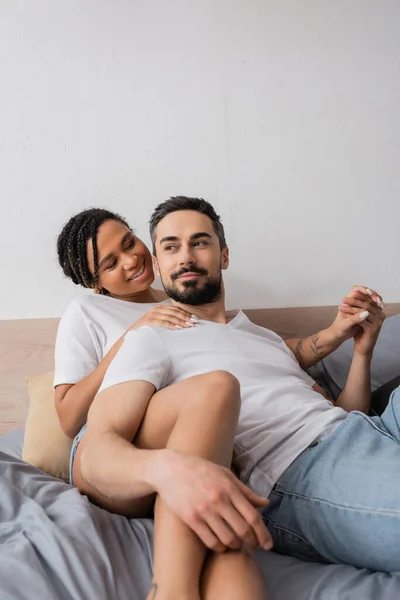 幸せな異人種間のカップルでホワイトTシャツ保持手ながら横たわっベッドに家 — ストック写真