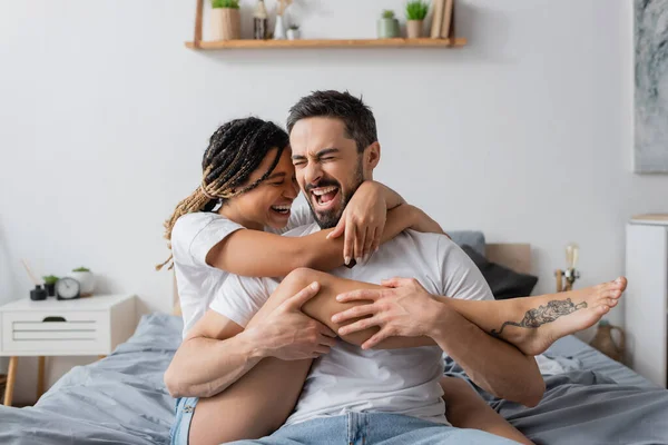 excited interracial couple in white t-shirts embracing while having fun in bedroom at home