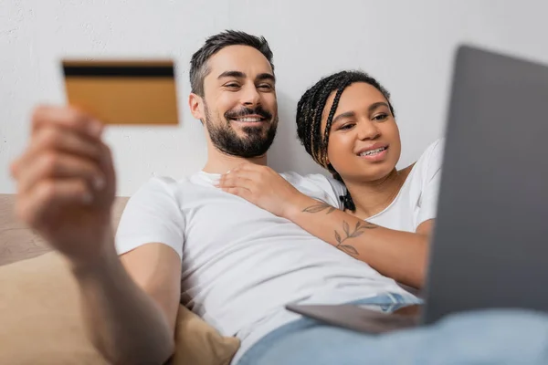 ラップトップとクレジットカードを持つ異人種間のカップルは 自宅のベッドルームでぼやけたフォアグラウンドでオンラインショッピング中に笑顔 — ストック写真