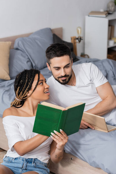 surprised and happy african american woman showing book to smiling boyfriend lying in bedroom at home