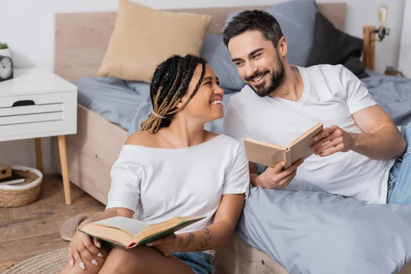 cheerful interracial couple in white t-shirts holding books and smiling at each other in bedroom at home