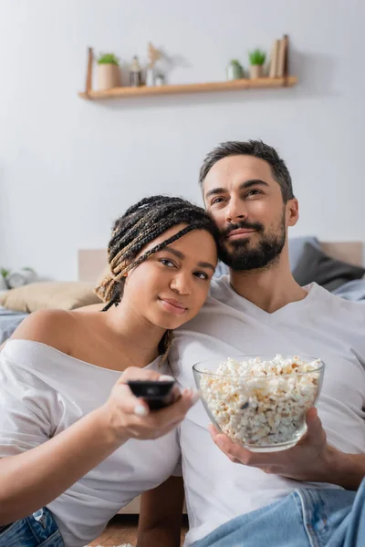 笑顔アフリカ系アメリカ人女性とともにテレビリモコンクリックテレビチャンネル近くひげを生やしたボーイフレンドとともにボールのポップコーンでベッドルームに自宅 — ストック写真