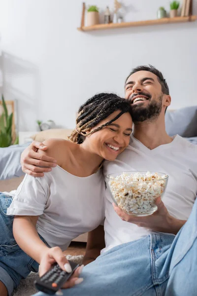 Lachender Bärtiger Mann Mit Popcorn Umarmt Lächelnde Afroamerikanerin Mit Fernbedienung — Stockfoto