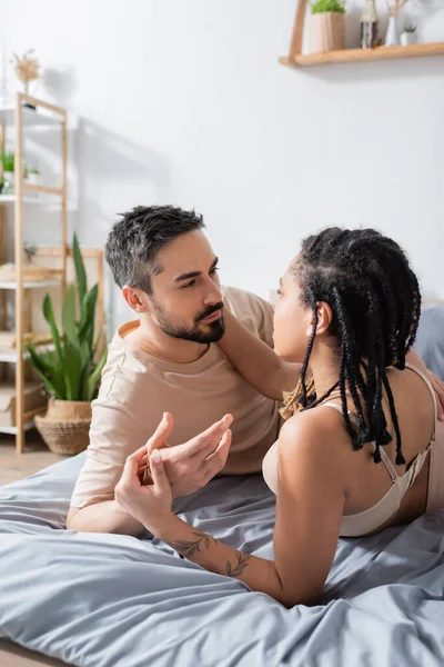 穿着胸罩的性感而纹身的非洲裔美国女人在家里的床上引诱着穿着T恤的留着胡子的男人 — 图库照片