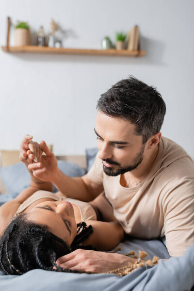 bearded man in t-shirt holding hand of passionate african american woman lying on bed at home