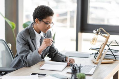 stylish african american businesswoman in grey blazer and eyeglasses sitting with pen near blank notebook and laptop in modern office clipart