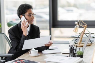 Gözlüklü şık afro-amerikan iş kadını akıllı telefondan konuşuyor ve evrakları ofisinde tutuyor.