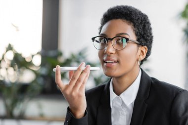 Siyah ceketli ve gözlüklü Afro-Amerikan iş kadını ofisteki akıllı telefondan sesli mesaj kaydediyor.