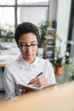 Beyaz bluzlu ve gözlüklü Afro-Amerikan yönetici ofiste deftere yazıyor.