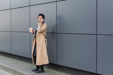 african american businesswoman in beige trench coat standing with takeaway drink and calling on smartphone near grey wall in city clipart