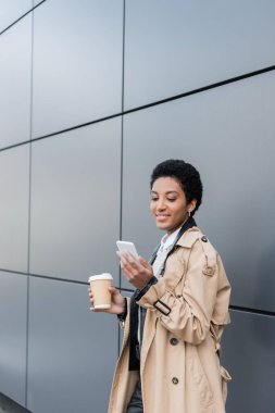 Bej renkli trençkotlu Amerikalı neşeli Afro-Amerikalı iş kadını elinde kağıt bardakta kahve tutuyor ve şehirdeki gri duvarın yanında akıllı telefondan sohbet ediyor.