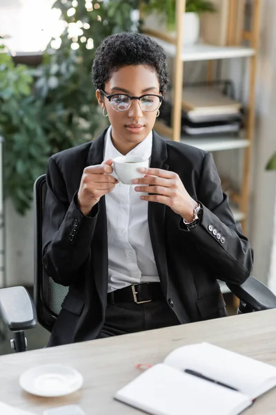 stock image african american businesswoman in stylish formal wear and eyeglasses holding coffee cup and looking at blank notebook in office